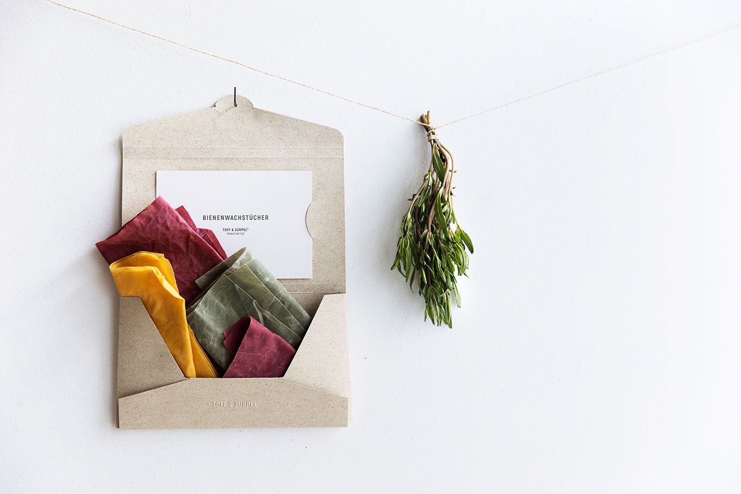Unsere wiederverwendbare Graspapier-Verpackung offen aufgehängt und gefüllt mit Bienenwachstücher in den Farben Grün, Rot und Gelb, daneben hängt ein Bündel Kräuter.