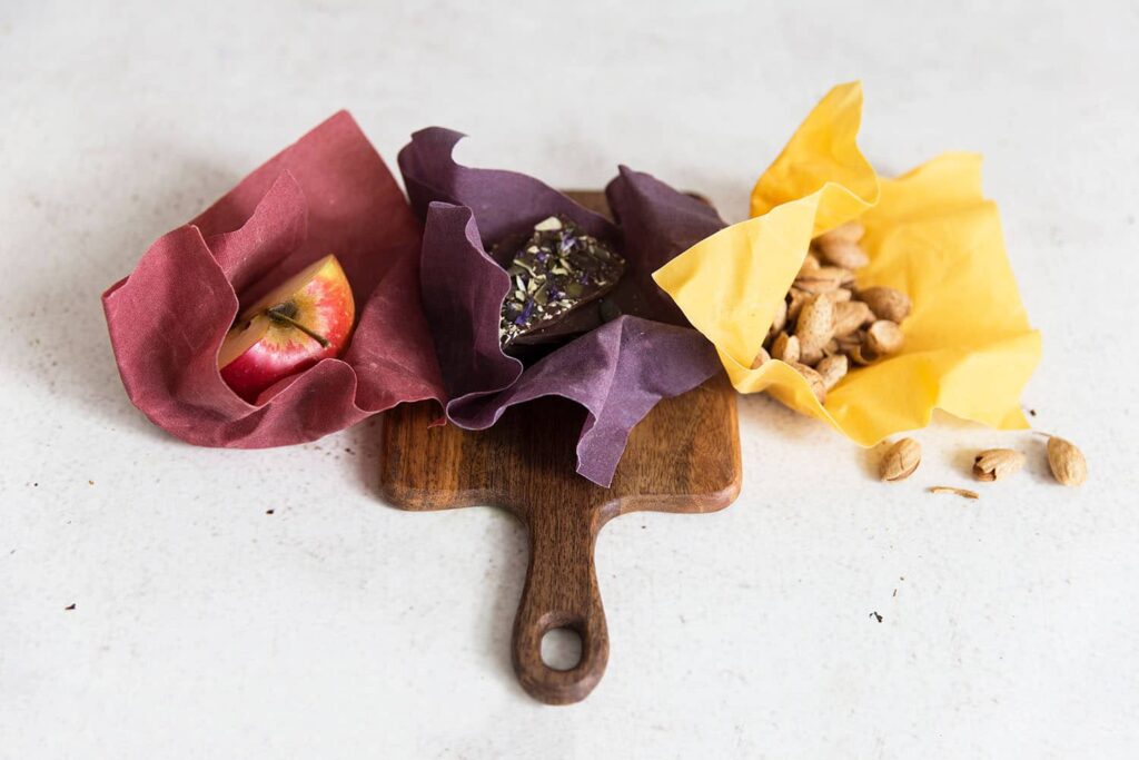 Three beeswax wraps in the colors red, purple and yellow lie on a board and cover nuts, a piece of an apple and a chocolate.