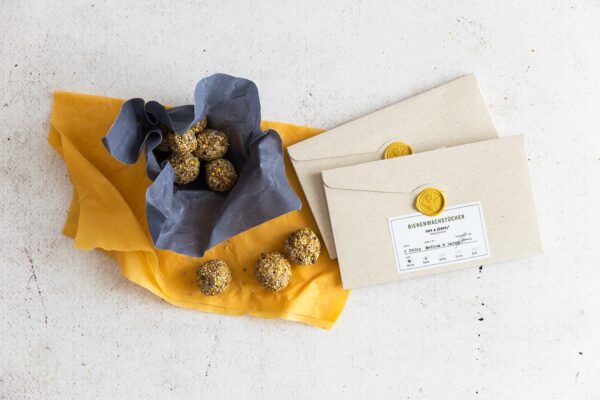 Two beeswax wraps in the colors yellow and blue filled with Energy Balls, next to them are two packaging for the beeswax wraps.