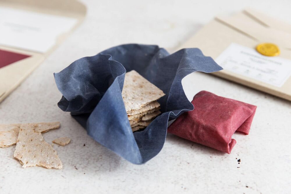 Two beeswax wraps in red and blue filled with crispbread, in the background are the product packaging for the beeswax cloths.