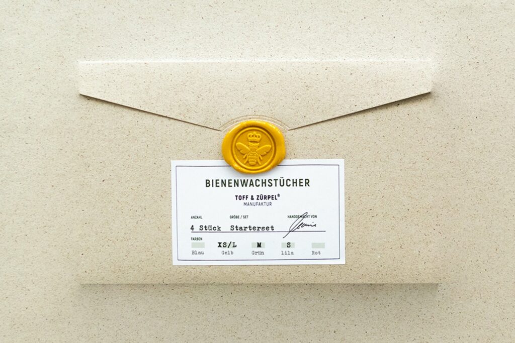 Die Verpackung aus Graspapier mit einem Bienen-Siegelstempel fÃ¼r das Bienenwachstücher Starterset in Gelb, Grün & Lila.
