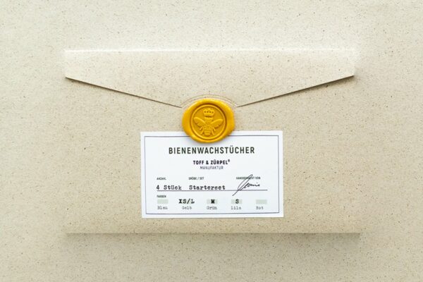 Die Verpackung aus Graspapier mit einem Bienen-Siegelstempel für das Bienenwachstücher Starterset in Gelb, Grün & Lila.